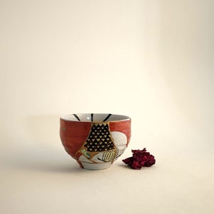 Può includere: Ciotola in ceramica rossa e bianca con una riparazione kintsugi unica. La ciotola presenta un motivo geometrico nero e oro. Un piccolo mucchio di fiori rossi secchi è accanto alla ciotola. Oggetto decorativo.