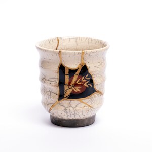 Può includere: Una tazza da tè in ceramica bianca con una glassa screpolata e dettagli dorati. La tazza ha un design nero e rosso di bambù e foglie.