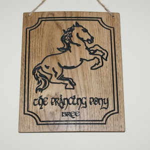 May include: Wooden sign featuring a carved black outline of a rearing horse. The sign reads "The Prancing Pony" and "Bree" in a stylized font. The rectangular sign has a decorative border and a jute string for hanging.