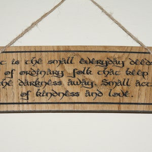 May include: Wooden sign with a quote in black script, hanging by a natural twine. The sign reads, "It is the small everyday deeds of ordinary folk that keep the darkness away. Small acts of kindness and love."