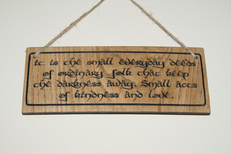 May include: Wooden sign with a quote in black script, hanging by a natural twine. The sign reads, "It is the small everyday deeds of ordinary folk that keep the darkness away. Small acts of kindness and love."