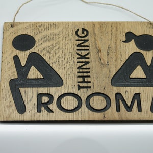 May include: A wooden sign with the text "Thinking Room" and two stick figures sitting on toilets. The sign is brown with black lettering and figures.