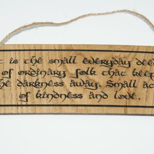 May include: Wooden sign with a natural wood grain finish and a jute rope hanger. The sign features black calligraphy text that reads, "It is the small everyday deeds of ordinary folk that keep the darkness away. Small acts of kindness and love."