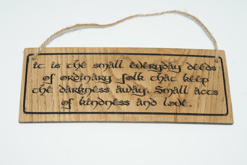 May include: Wooden sign with a natural wood grain finish and a jute rope for hanging. The sign features black calligraphy text that reads, "It is the small everyday deeds of ordinary folk that keep the darkness away. Small acts of kindness and love."