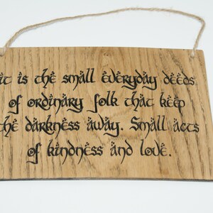 May include: Wooden sign with the quote "It is the small everyday deeds of ordinary folk that keep the darkness away. Small acts of kindness and love." The text is in black calligraphy on a light brown wooden plaque.