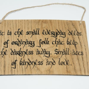 May include: Wooden sign with the quote "It is the small everyday deeds of ordinary folk that keep the darkness away. Small acts of kindness and love." The text is in black calligraphy on a light brown wooden plaque.