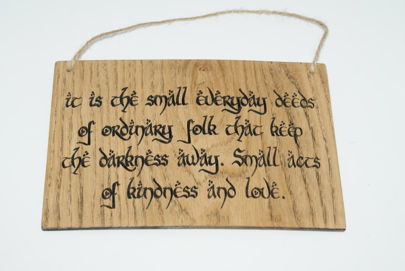 May include: Wooden sign with the quote "It is the small everyday deeds of ordinary folk that keep the darkness away. Small acts of kindness and love." The text is in black calligraphy on a light brown wooden plaque.