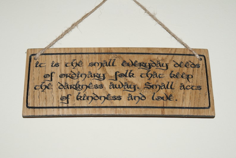 May include: Wooden rectangular sign with a quote in black calligraphy: "It is the small everyday deeds of ordinary folk that keep the darkness away. Small acts of kindness and love." The sign hangs by a twine.