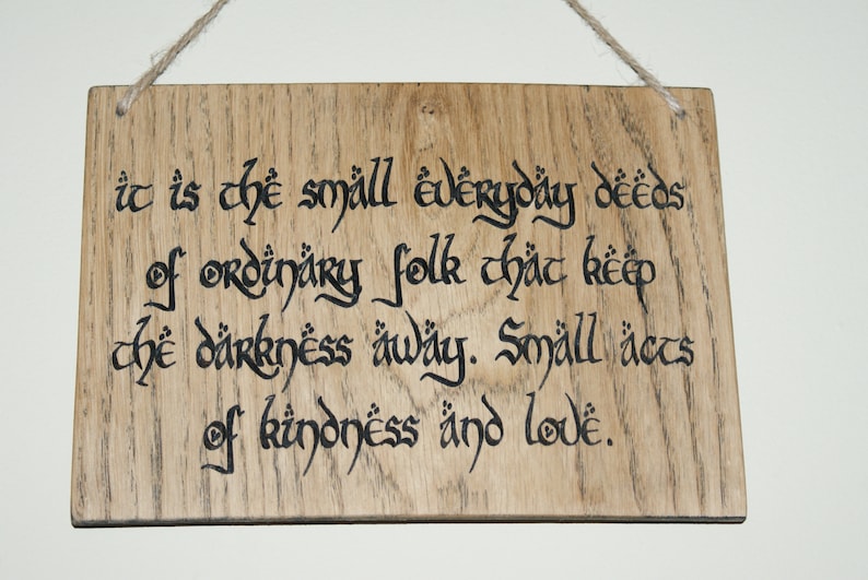 May include: Wooden sign with the quote "It is the small everyday deeds of ordinary folk that keep the darkness away. Small acts of kindness and love." The text is in black calligraphy on a light brown wood.