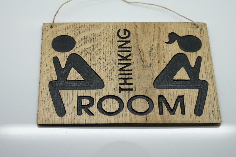 May include: A wooden sign with the text "Thinking Room" and two stick figures sitting on toilets. The sign is brown with black lettering and figures.