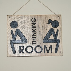 May include: A wooden sign with the text "Thinking Room" and two stick figures, one with a ponytail, sitting on a toilet.