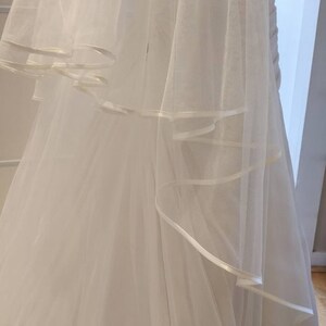May include: A white tulle wedding veil with a satin ribbon trim. The veil is layered and has a flowing design.