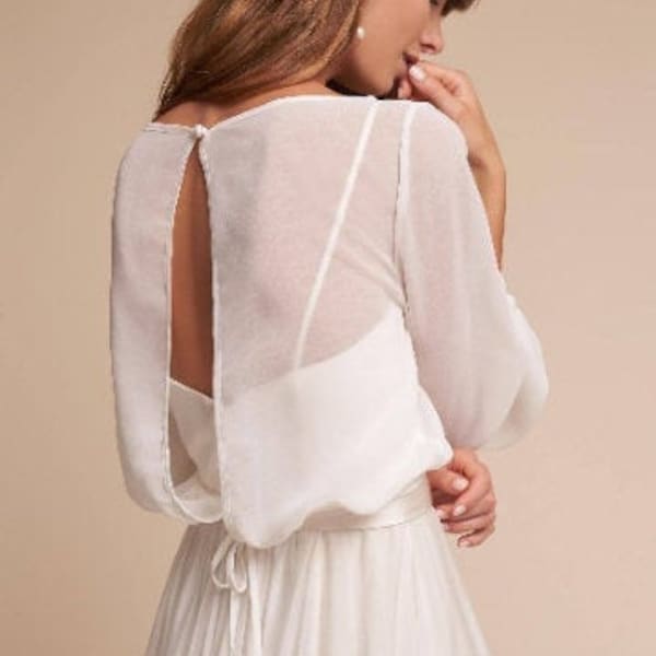May include: White wedding dress with a sheer, long-sleeved top and a flowing skirt. The top has a keyhole back and a tie at the waist.