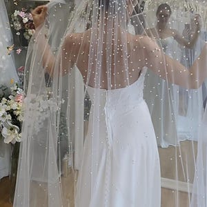 May include: A white bridal gown with a strapless bodice and a flowing skirt. A sheer veil, embellished with scattered pearls, is draped over the shoulders and back. The dress is simple and elegant, suitable for a wedding.