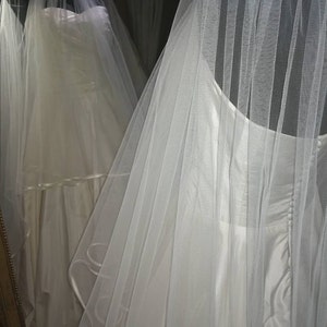 May include: A white wedding dress with a tulle veil. The dress has a strapless bodice and a flowing skirt. The veil is layered and has a scalloped edge.