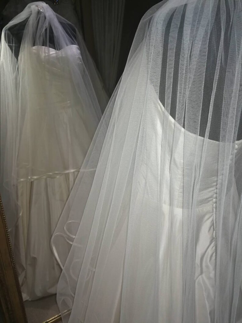 May include: A white wedding dress with a tulle veil. The dress has a strapless bodice and a flowing skirt. The veil is layered and has a scalloped edge.