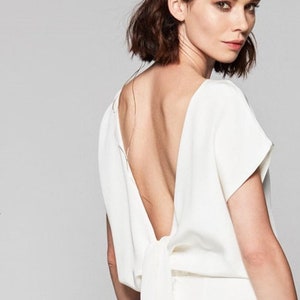 Könnte beinhalten: Ein weißes ärmelloses Kleid mit tiefem Rücken und einem Schleifen-Detail. Das Kleid ist aus einem weichen, fließenden Stoff.