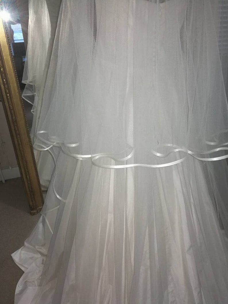 May include: A white wedding dress with a tulle skirt and a white satin ribbon trim. The dress has a long train and a fitted bodice.