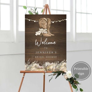Pode incluir: Placa de boas-vindas imprimível para um chá de panela com um fundo de madeira rústico. A placa apresenta um par de botas de cowboy marrons com um laço de renda branco e uma corda de luzes. O texto na placa diz "Bem-vinda ao chá de panela da Jennifer".