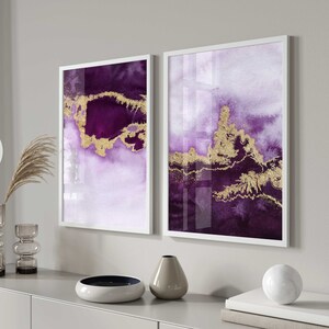 May include: Two framed abstract art prints with white frames. The prints feature a purple and gold watercolor design. The prints are hanging on a white wall above a gray dresser with a vase, a bowl, and a decorative ball on top.