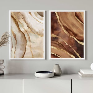 May include: Two framed abstract paintings with a white background and brown and gold abstract shapes. The paintings are hanging on a white wall above a white cabinet.