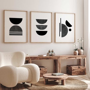 May include: Three framed black and white abstract geometric prints with a white background hanging on a white wall above a wooden table with a white armchair and a round wooden coffee table.