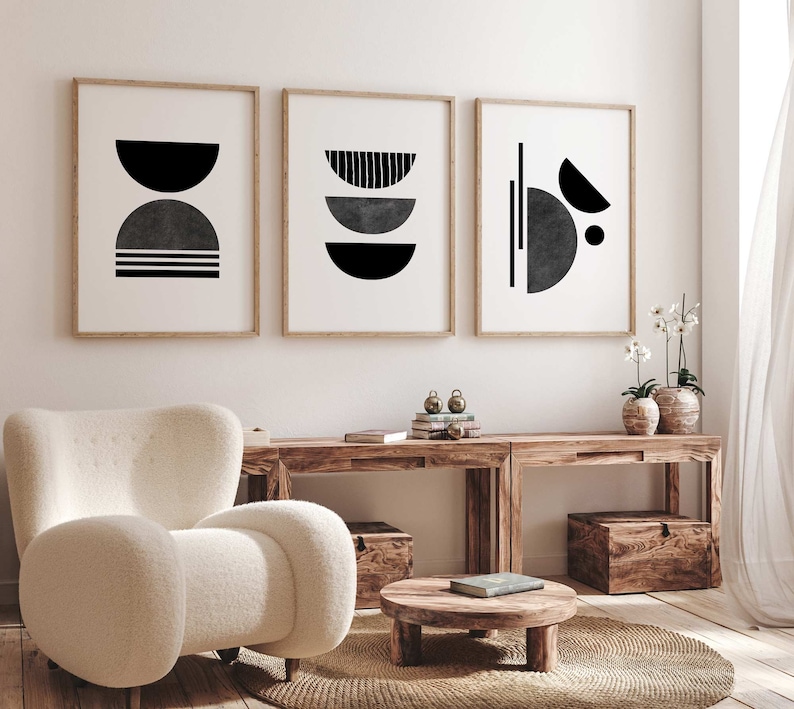May include: Three framed black and white abstract geometric prints with a white background hanging on a white wall above a wooden table with a white armchair and a round wooden coffee table.