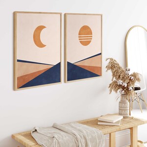 May include: Two framed prints with abstract geometric designs in shades of peach, brown, and blue. The prints feature a crescent moon and a sun with horizontal lines. The prints are hanging on a white wall above a wooden bench with a woven seat and a white throw blanket.