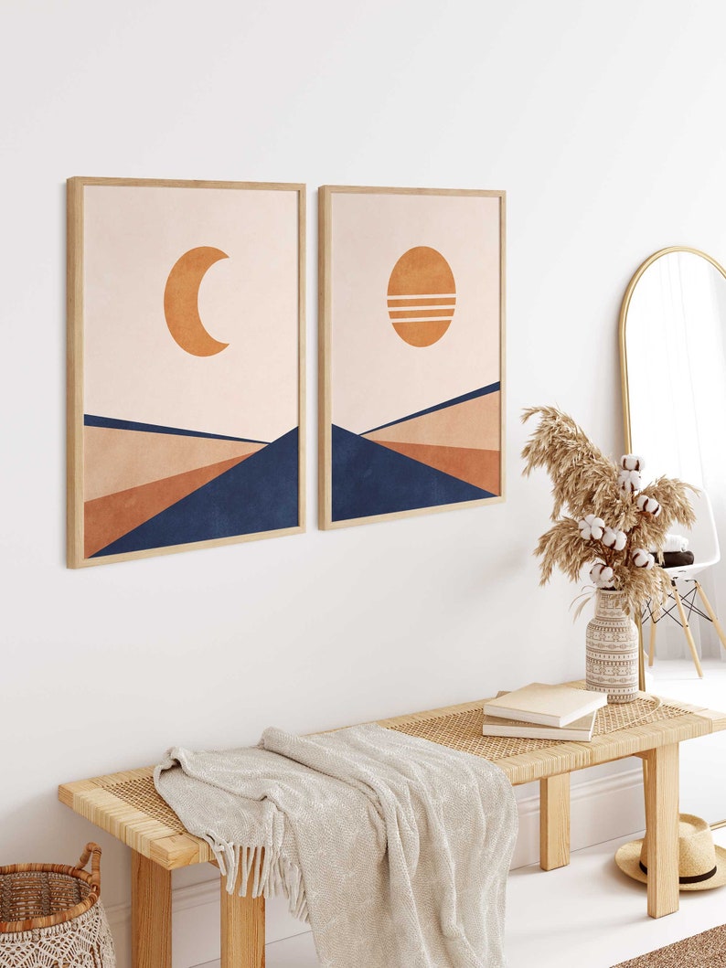 May include: Two framed prints with abstract geometric designs in shades of peach, brown, and blue. The prints feature a crescent moon and a sun with horizontal lines. The prints are hanging on a white wall above a wooden bench with a woven seat and a white throw blanket.