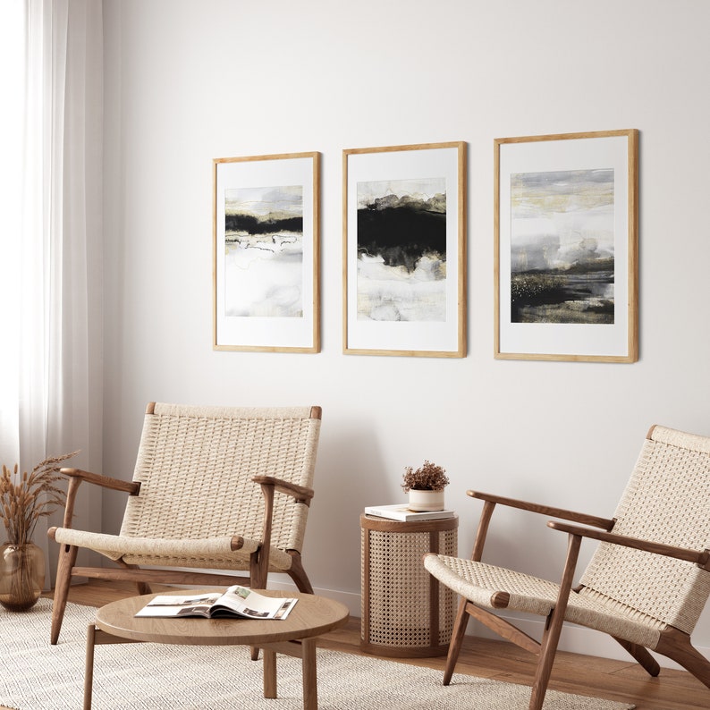 May include: Three framed abstract landscape paintings in black, white, and gold tones hang on a white wall above two woven chairs and a woven side table.