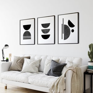 May include: Three black and white abstract geometric art prints with a minimalist design. The prints are framed in black and hung on a white wall above a white couch.