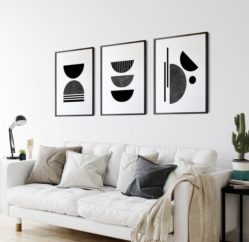 May include: Three black and white abstract geometric art prints with a minimalist design. The prints are framed in black and hung on a white wall above a white couch.