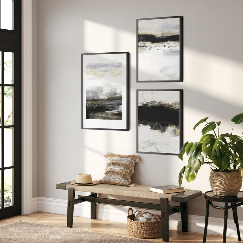 May include: Three black framed abstract art prints with white, gray, and black paint. The prints are hanging on a white wall above a wooden bench with a woven cushion and a straw hat.