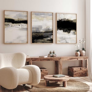 May include: Three abstract landscape paintings in wooden frames, with muted tones of gray, white, black, and gold. The paintings are hanging on a white wall above a wooden console table with two potted plants.