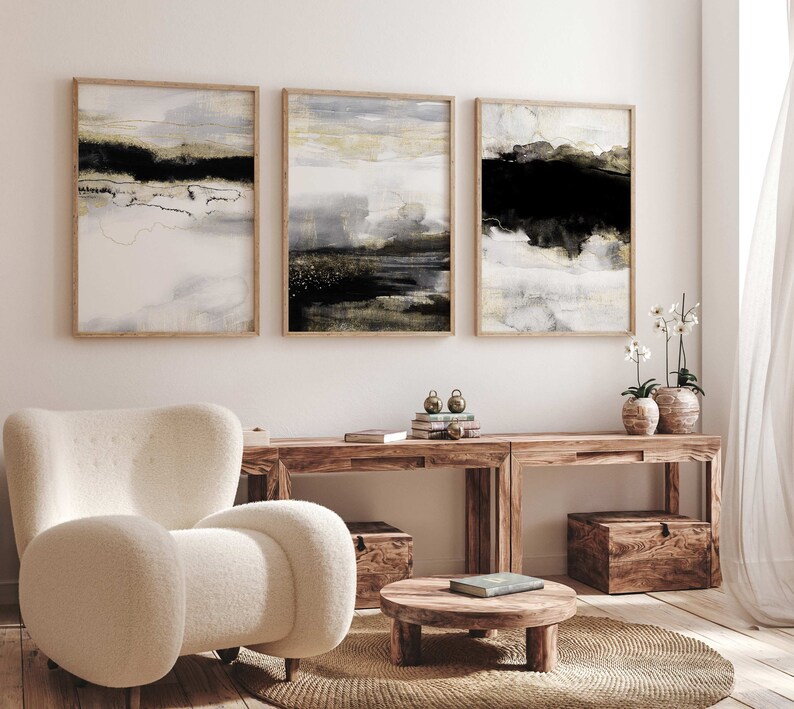 May include: Three abstract landscape paintings in wooden frames, with muted tones of gray, white, black, and gold. The paintings are hanging on a white wall above a wooden console table with two potted plants.