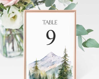 Op de afbeelding: Een witte tafelnummerkaart met een gouden frame. De kaart heeft de tekst "TABLE 9" en een aquarel-illustratie van een bergketen met bomen.