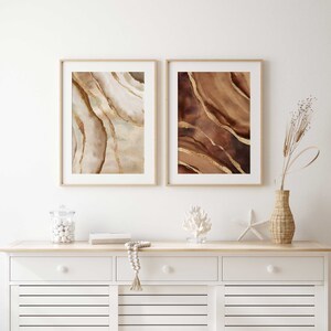 Peut inclure: Deux impressions d'art abstrait encadr&eacute;es avec des motifs d'aquarelle brun et or. Les impressions sont accroch&eacute;es &agrave; un mur blanc au-dessus d'une commode blanche &agrave; deux tiroirs.