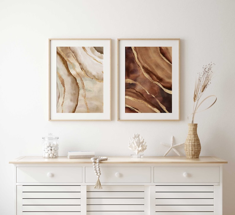 Peut inclure: Deux impressions d'art abstrait encadr&eacute;es avec des motifs d'aquarelle brun et or. Les impressions sont accroch&eacute;es &agrave; un mur blanc au-dessus d'une commode blanche &agrave; deux tiroirs.
