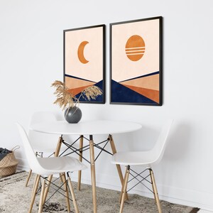 May include: Two abstract art prints with a minimalist design. The prints feature a crescent moon and a sun with a geometric pattern on a peach background. The prints are framed in black and have a navy blue geometric pattern at the bottom.