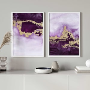 Puede incluir: Dos pinturas abstractas de acuarela enmarcadas con detalles dorados. Las pinturas son en tonos de morado y blanco. Las pinturas est&aacute;n colgadas en una pared blanca sobre un estante gris.