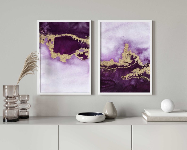 Puede incluir: Dos pinturas abstractas de acuarela enmarcadas con detalles dorados. Las pinturas son en tonos de morado y blanco. Las pinturas est&aacute;n colgadas en una pared blanca sobre un estante gris.