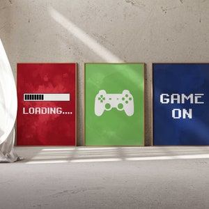 Puede incluir: Tres impresiones enmarcadas con un tema de juegos. La primera impresi&oacute;n es roja con una barra de carga blanca y el texto "CARGANDO..." La segunda impresi&oacute;n es verde con un icono de mando de videojuego blanco. La tercera impresi&oacute;n es azul con el texto "GAME ON" en blanco.