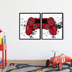 May include: Two framed prints featuring red video game controllers against a white background with black paint splatter accents. The prints are displayed on a white wall above a gray rug with a race track design. A red toy car and a red side table with a lamp are in the foreground.