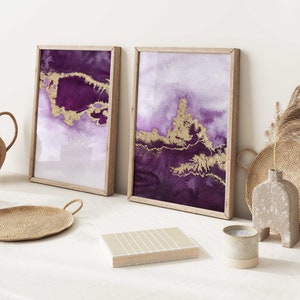 Puede incluir: Dos pinturas abstractas de acuarela enmarcadas con detalles dorados. Las pinturas son en tonos de morado y blanco. Las pinturas est&aacute;n colgadas en una pared blanca.