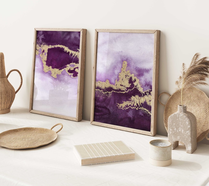 Puede incluir: Dos pinturas abstractas de acuarela enmarcadas con detalles dorados. Las pinturas son en tonos de morado y blanco. Las pinturas est&aacute;n colgadas en una pared blanca.