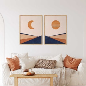 May include: Two abstract landscape prints featuring a crescent moon and a sun with two lines across it. The prints are framed in a light wood frame and are hanging on a white wall above a white sofa with cushions.