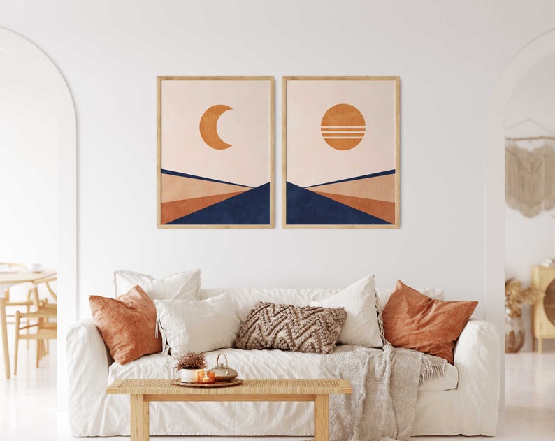May include: Two abstract landscape prints featuring a crescent moon and a sun with two lines across it. The prints are framed in a light wood frame and are hanging on a white wall above a white sofa with cushions.