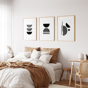 May include: Three black and white abstract geometric prints in wooden frames hanging on a white wall above a bed with a brown blanket and white bedding.