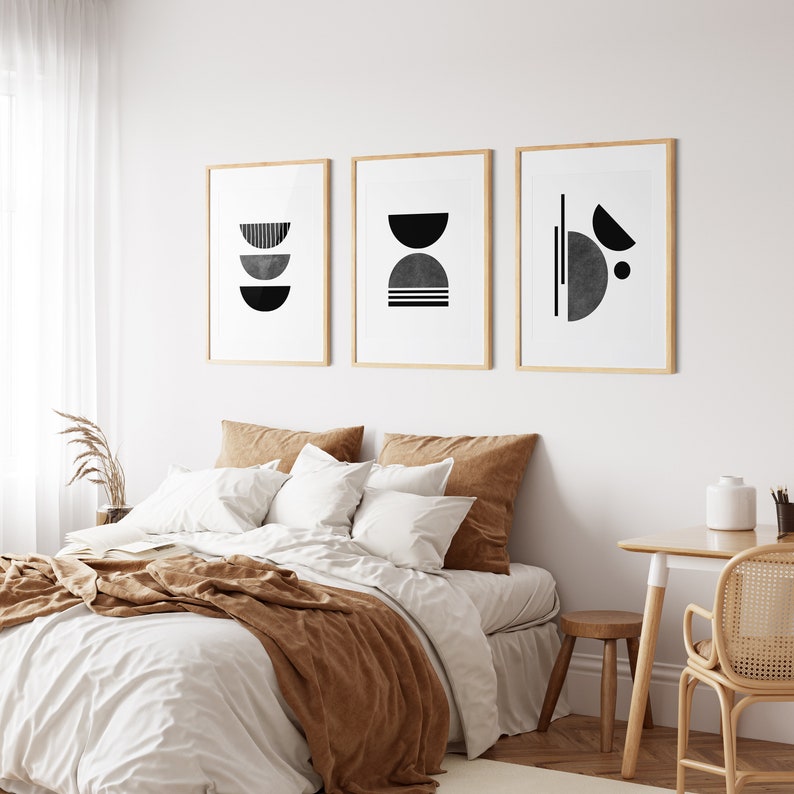 May include: Three black and white abstract geometric prints in wooden frames hanging on a white wall above a bed with a brown blanket and white bedding.