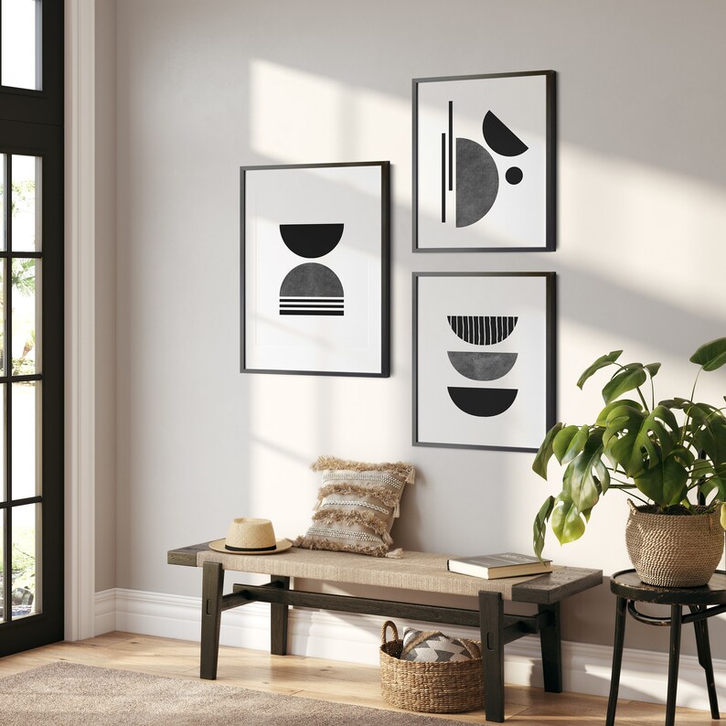 May include: Three black framed prints with abstract geometric designs in shades of gray and black. The prints are hung on a white wall above a wooden bench with a woven cushion and a straw hat.
